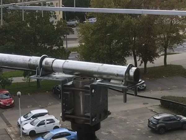 Zbliżenie dodatkowego mocowania: gruba rura podpory w masywnej obejmie na czarnym stalowym wysięgniku z balkonu, nad nią bom anteny
