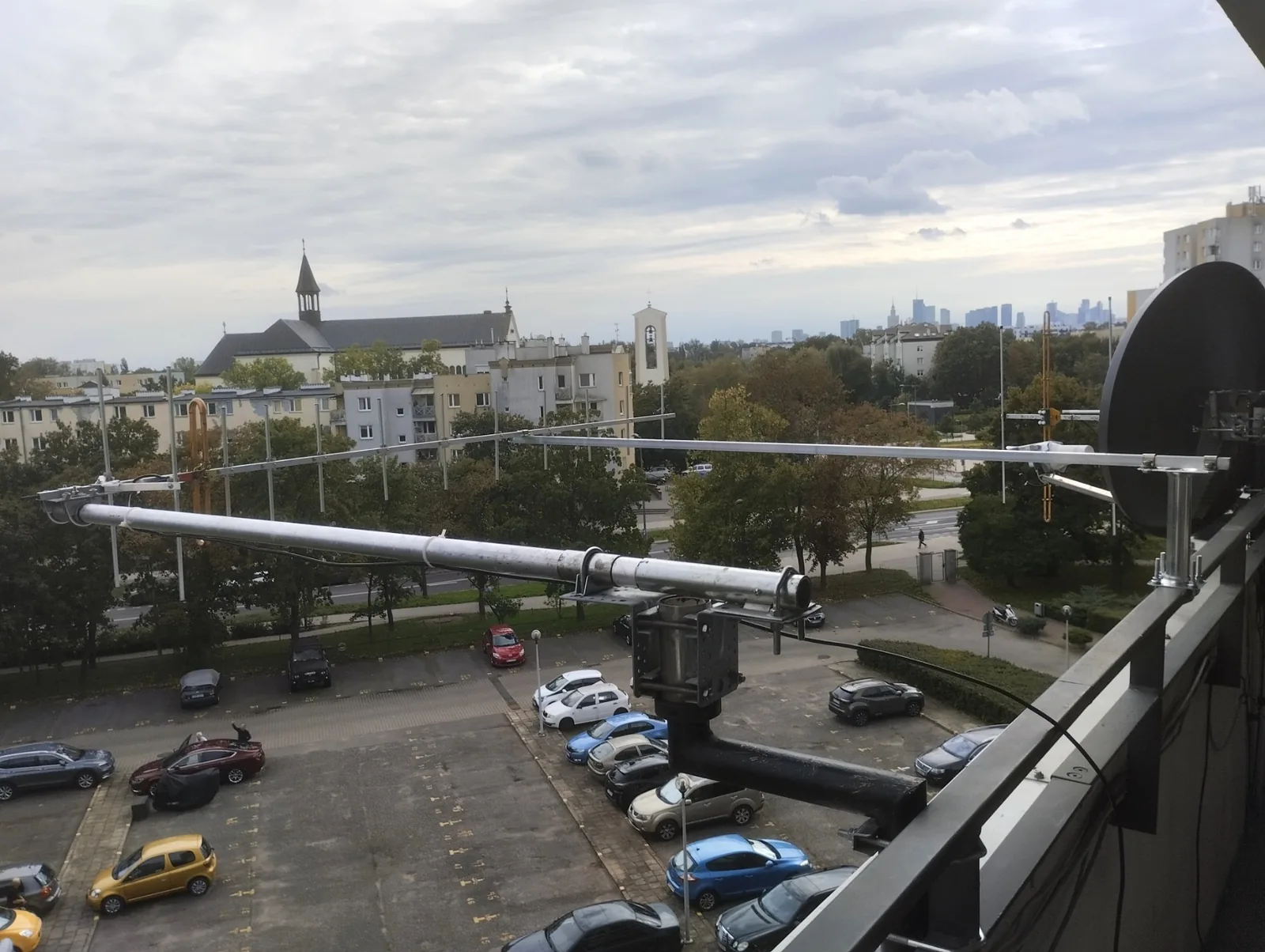 Długa antena Yagi na 70 cm z pionowymi elementami na balkonie wysokiego piętra; pod bomem druga rura podpierająca, w tle osiedle, kościół i wieżowce centrum Warszawy