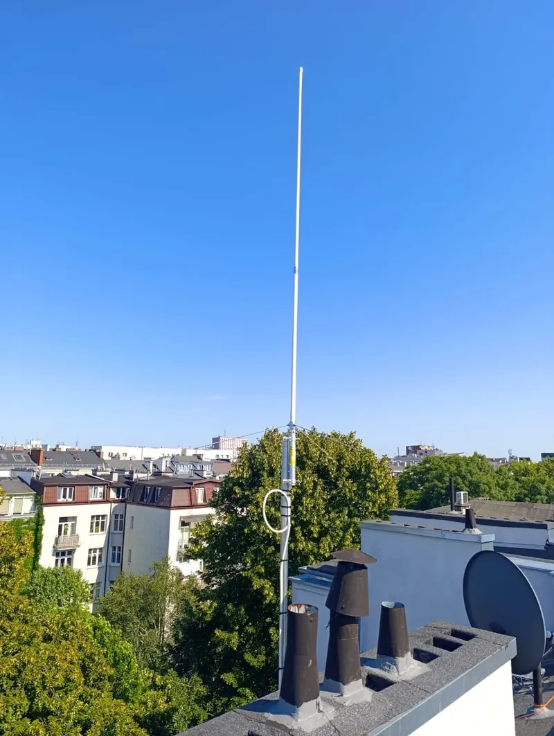 Biała antena pionowa Diamond X-200N na maszcie ponad kominami kamienicy, w tle dachy Mokotowa, korony drzew i bezchmurne niebo