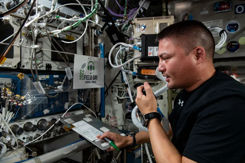 Astronauta w module stacji kosmicznej mówi do mikrofonu radiostacji amatorskiej, trzymając notatki, wokół kable i aparatura.