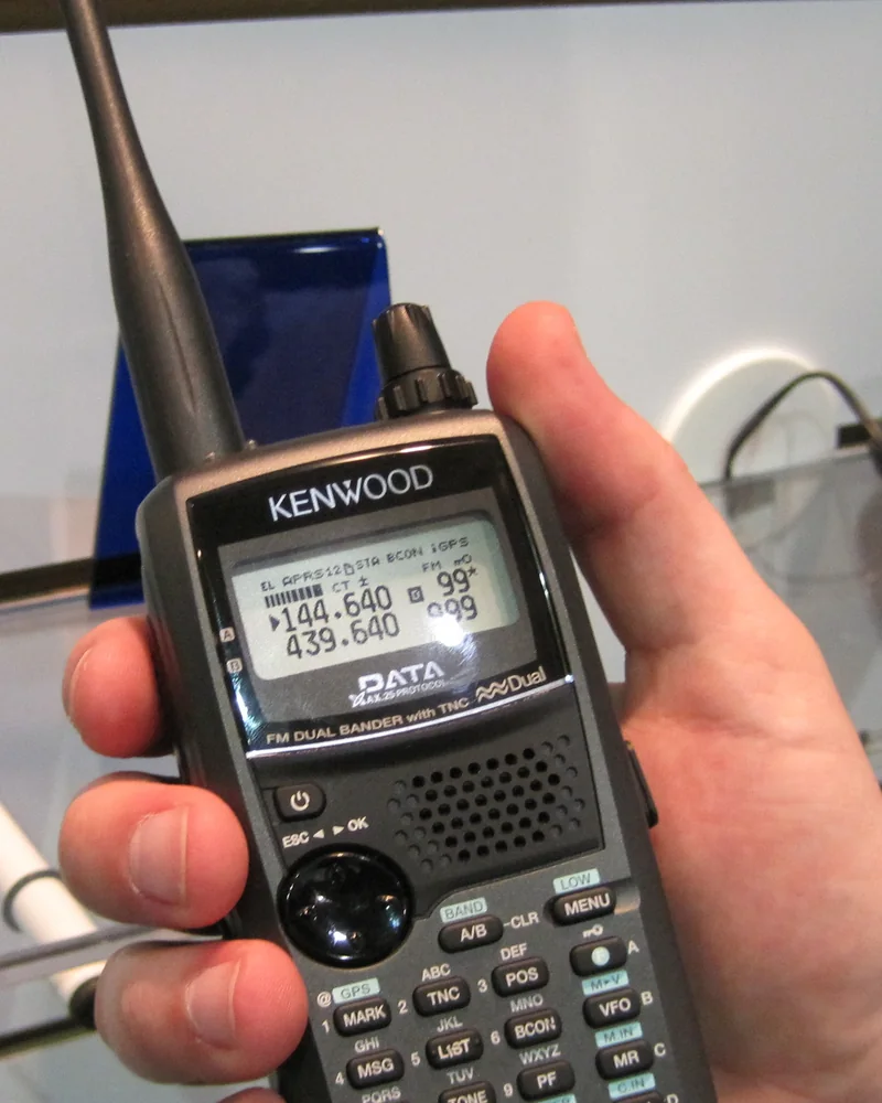 Dłoń trzyma ręczny radiotelefon dwupasmowy, na wyświetlaczu widać napisy APRS i GPS oraz częstotliwości 144.640 i 439.640.