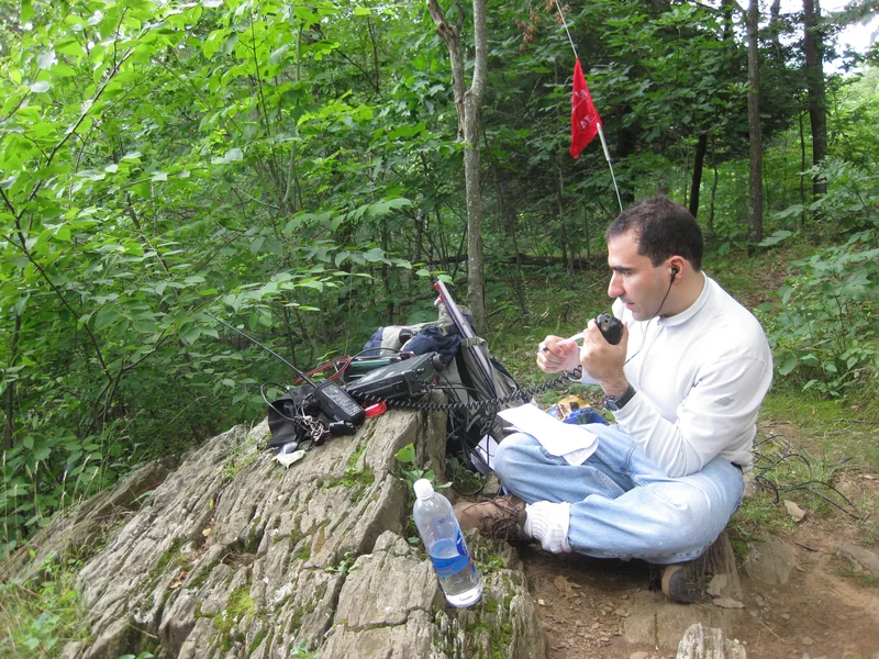 Mężczyzna siedzi na skale w lesie obok sprzętu radiowego i mówi do mikrofonu ręcznego, nad nim antena z czerwoną flagą.
