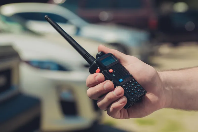 Dłoń trzymająca dwupasmowy radiotelefon ręczny z wyciągniętą anteną i klawiaturą, w tle rozmyte samochody na parkingu