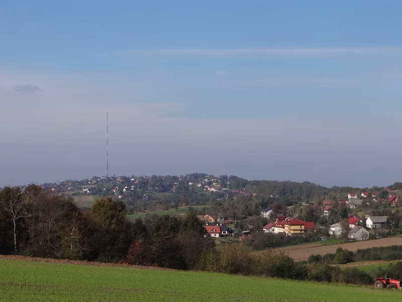 Wzgórze z cienkim masztem ponad zabudową wsi, na pierwszym planie pole
