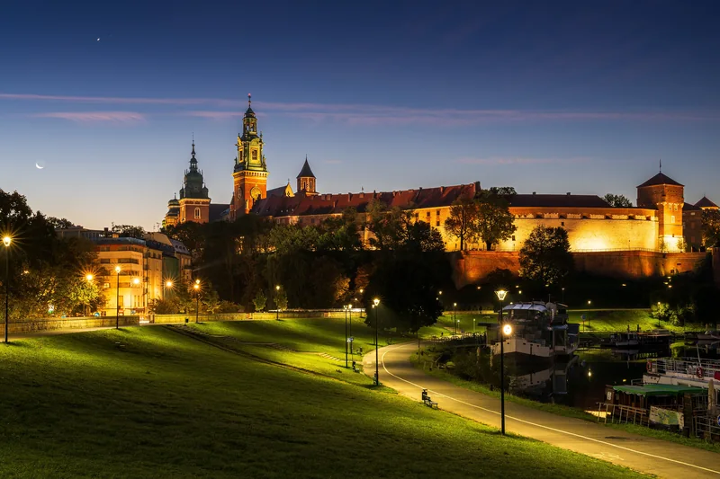 Oświetlony zamek na Wawelu o zmierzchu, na pierwszym planie bulwary i latarnie