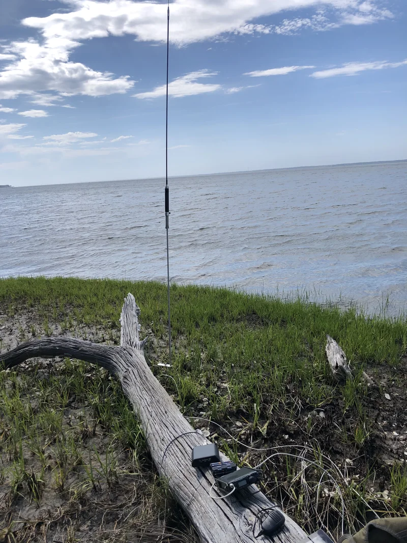 Pionowa antena na maszcie nad brzegiem jeziora, na pniu w trawie leży małe radio z akumulatorem