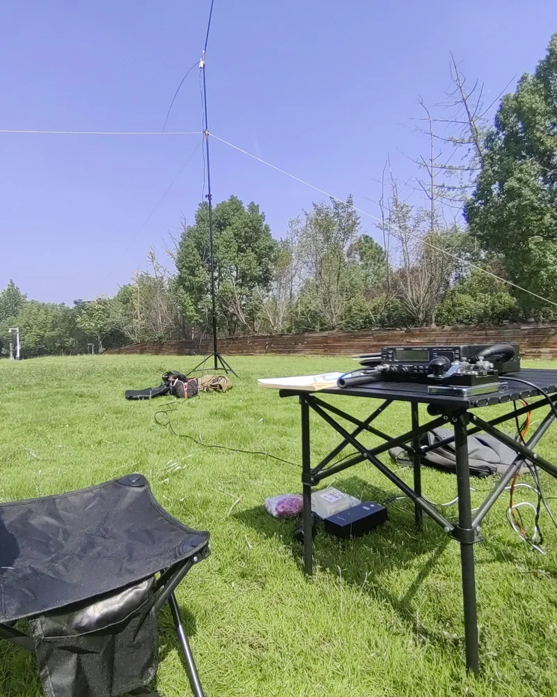 Składany stolik z radiem i klawiszem telegraficznym na trawiastej polanie, obok krzesełko turystyczne, w tle maszt z rozpiętą anteną drutową i drzewa