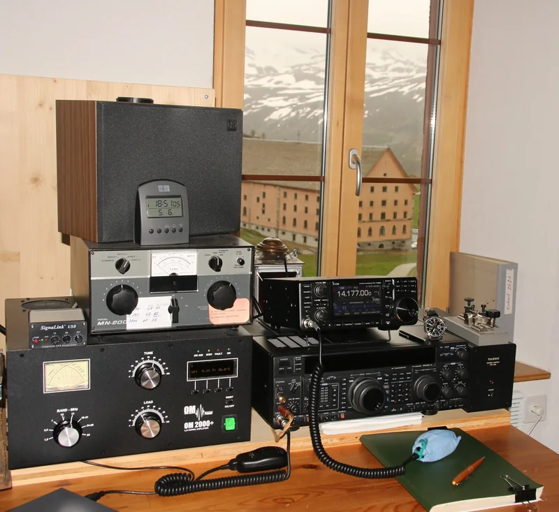 Stacja krótkofalarska przy oknie z widokiem na góry: transceivery Icom IC-7300 i Yaesu FT-1000MP Mark-V, wzmacniacz, klucz telegraficzny, a na biurku zielony zeszyt logu z długopisem