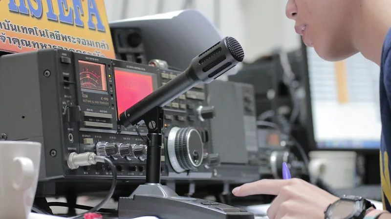 Krótkofalowiec mówi do mikrofonu stołowego przed transceiverem Icom IC-970, w dłoni trzyma długopis do notowania łączności