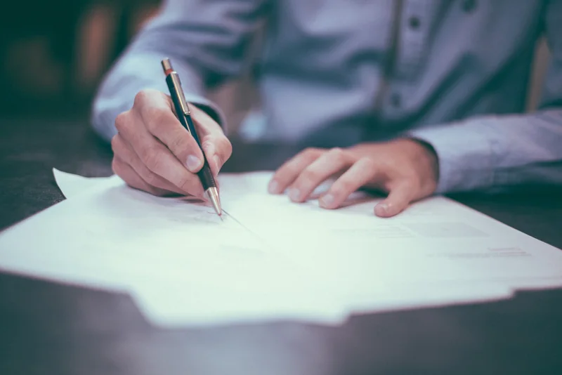 Dłoń trzymająca długopis wypełnia kartkę papieru leżącą na stole