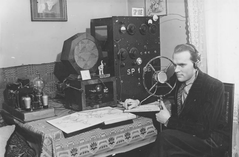 Czarno-białe zdjęcie z 1939 roku: Michał Wysokiński SP1QE w słuchawkach przy lampowym nadajniku z napisem SP1QE i mikrofonie w ramie, notuje ołówkiem w dzienniku łączności, obok otwarty atlas
