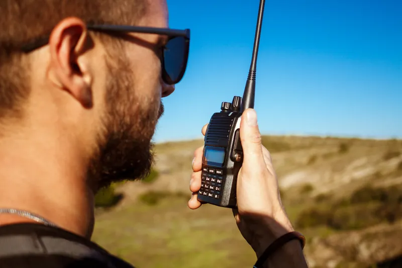 Zbliżenie: mężczyzna w okularach przeciwsłonecznych trzyma ręczny radiotelefon z anteną blisko twarzy, w tle wzgórza i błękitne niebo