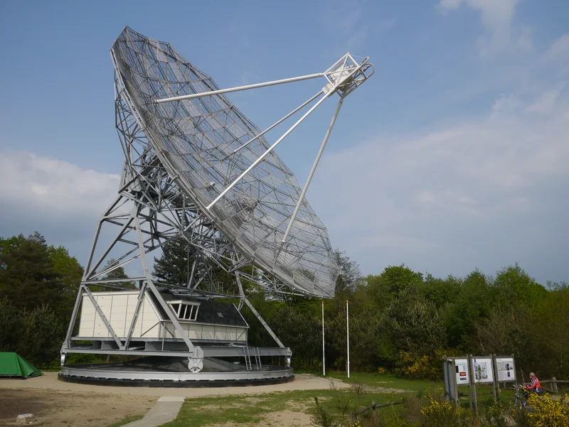 Duża ażurowa czasza radioteleskopu na stalowej konstrukcji z budynkiem u podstawy, otoczona drzewami.