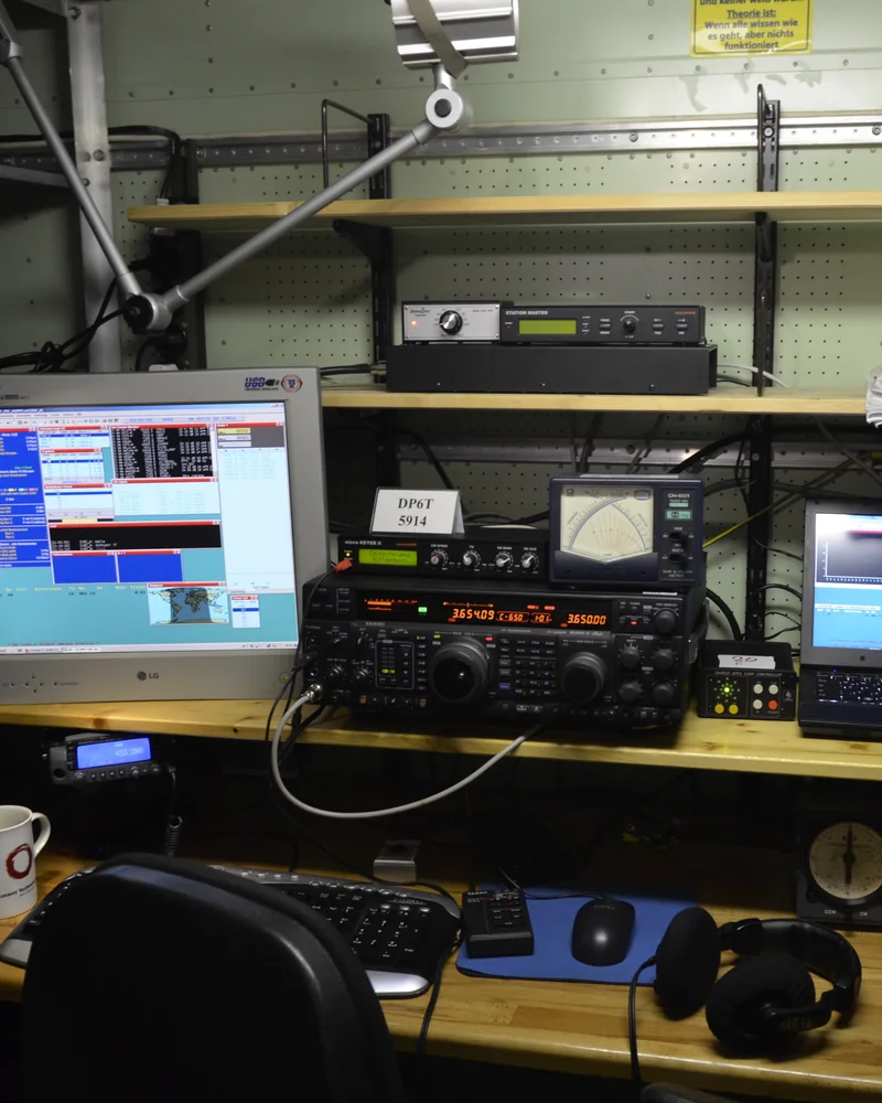 Stanowisko krótkofalarskie: transceiver na półce, monitor i laptop z programami do łączności, a na blacie klawiatura i słuchawki.