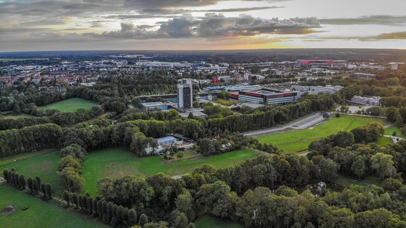 Widok z lotu ptaka na zielony kampus uniwersytecki z budynkami wśród drzew i miastem w tle o zachodzie słońca.