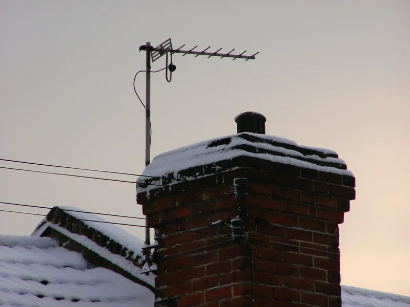 Antena Yagi na maszcie przymocowanym do ceglanego komina, dachy i komin przysypane śniegiem o zmierzchu